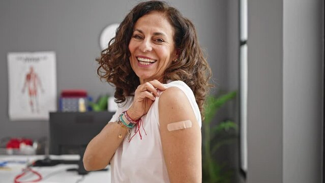 Middle age hispanic woman smiling confident standing with band aid on arm at clinic