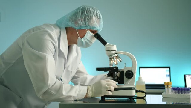 Male medical worker, wearing lab coat, medical hat, mask and gloves researches sample on microscope in lab in vet clinic. Laptop, tablet, test tubes. High quality 4k footage