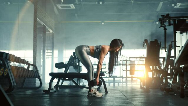 Process of performing dumbbell Romanian deadlift by fit beautiful dark-haired woman in gym. Overview of fully equipped gym. Misty background with sun rays. High quality 4k footage