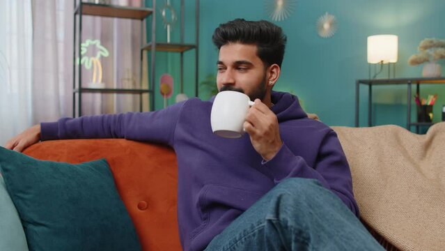 Handsome smiling indian man drinking a cup of warm coffee or herbal tea at home living room apartment. Break time. Hindu guy enjoying comfortable relaxing sitting on couch in the morning. Lifestyles