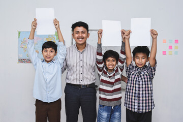 Obraz premium Smiling diverse student raising blank white paper and standing in the classroom with teacher . Copy space and mock up. 