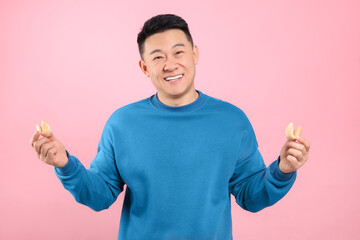 Fototapeta premium Happy asian man holding tasty fortune cookies with predictions on pink background