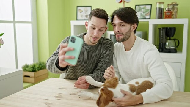 Two men couple taking selfie picture by smartphone sitting on table with dog at dinning room