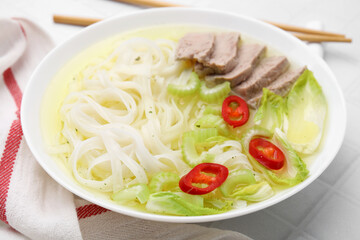 Bowl of delicious rice noodle soup with celery and meat on white tiled table, closeup