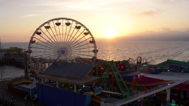 Aerial Panning Shot Of Ferris Wheel At Santa Monica Pier Against Sky, Drone Flying Over Beach At Sunset - Los Angeles, California