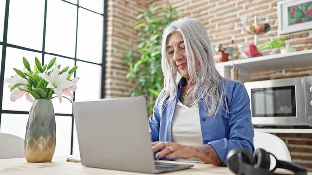 Middle age grey-haired woman using laptop sitting on table at dinning room