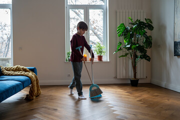 Teen boy doing chores cleaning floor in living room sweeping trash with broom to scoop. Teenager helping with household duties. Tidying up house involving children in family to maintain order at home.