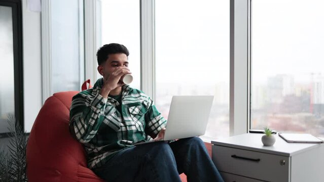 Young indian man in casual outfit and white sneakers sitting on orange bean bag chair and working on laptop in comfortable office. Focused worker holding cup of hot coffee and typing on keyboard.