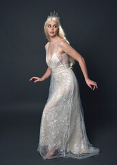 Full length portrait of beautiful women with long blonde hair, wearing fantasy  princess crown and elegant white ball gown, standing pose with hand gesture. Isolated on dark grey studio background.