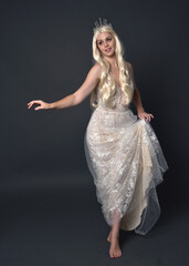 Fototapeta premium Full length portrait of beautiful women with long blonde hair, wearing fantasy princess crown and elegant white ball gown, standing pose with hand gesture. Isolated on dark grey studio background.