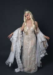 Full length portrait of beautiful women with long blonde hair, wearing fantasy  princess crown and elegant white ball gown, standing pose with hand gesture. Isolated on dark grey studio background. © faestock