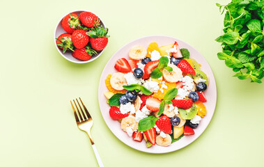 Summer fruit and berry salad with strawberries, blueberries, banana, soft cheese and mint leaves, green background, top view