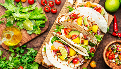 Taco party. Corn tortillas with grilled chicken fillet, salsa sauce, mango, cilantro and red onion on rustic wooden cutting board. Wood table background, top view