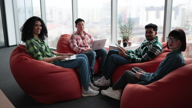 Diverse male and female adults in casual clothes posing on terracotta soft seats with digital gadgets. Multiethnic collaborators planning presentation for colleagues in shared workspace.
