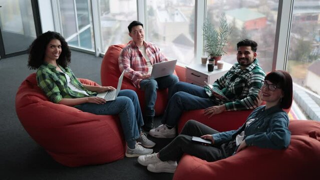 Top view of diverse male and female adults in casual clothes posing on terracotta soft seats with digital gadgets. Multiethnic collaborators planning presentation for colleagues in shared workspace.
