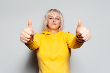 Studio portrait of happy blonde woman showing thumbs up on white background, wearing yellow sweater.