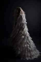 fantasy portrait of beautiful female model with long blond hair wearing otherworldly  white feathered cloak costume and headdress, isolated on dark studio background.