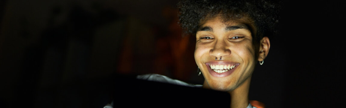 Portrait of joyful young guy smiling while using tablet pc at night, sitting in dark room at home
