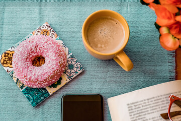 Break concept. Pink glazed donut on breakfast table, cup of coffee, book and mobile phone