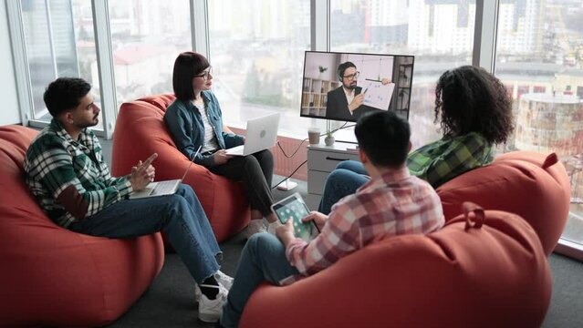 Focused diverse employees making video call on display while sitting with laptops and tablet on soft poufs in cozy workplace. Young self-employed workers taking part in online briefing in office.