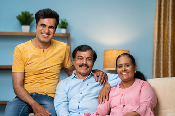 portrait shot of Happy smiling indian senior couple with adult son looking at camera while sitting on sofa at home - concept of family bonding, spending time and generation