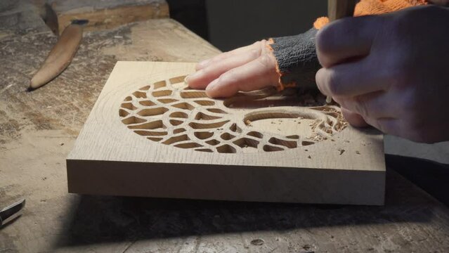 Close up hands of a wood carver using a small chisel to make a relief carving of a tree in circle shape from a flat square piece of wood, shot in slow motion