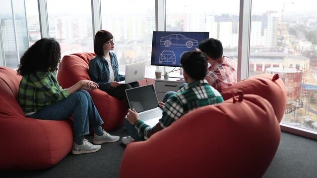 Back view of diverse male and female coworkers examining digital display while staying in open office with terracotta beanbags. Auto designers improving vehicle system using computer programs.
