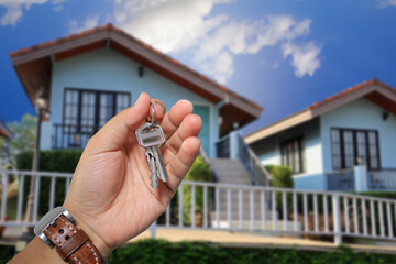 Male hand holding a key chain and a house in the background represents buying a house or investing...