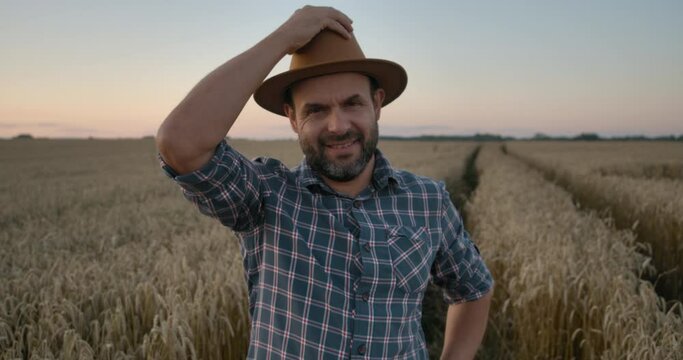 Portrait Farmer Bearded Man With Hat Standing in Wheat Field. Farm Worker. Sunset Sky. Portrait Caucasian Farmer Man in Plaid Shirt in Hat Looking at Camera. Farmland Sunset Landscape Agriculture. 