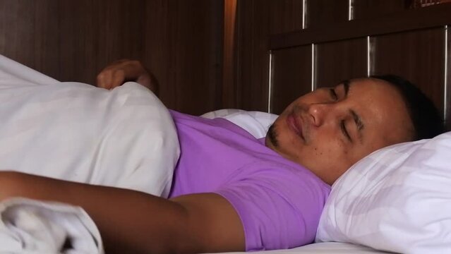 An asian man getting ready for bed but with a restless expression in his bed, HD video