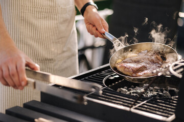 Male chef preparing T-bone steak on carbon steel frying pan outdoor on grill. BBQ concept. Close-up