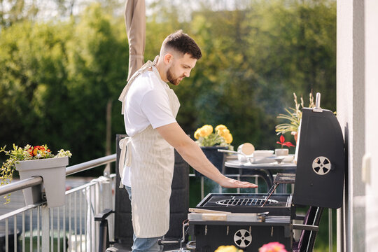 Side view of handsome man try how hot is BBQ grill. Man in apron make dinner outdoor