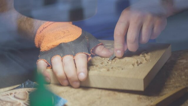 Close up hands of a wood carver using a small chisel to make a relief carving of a tree in circle shape from a flat square piece of wood, shot through a glass window showing outdoor reflection