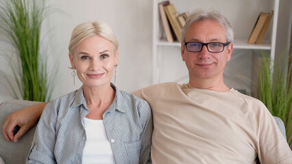 Middle-aged couple. Positive day. Family shooting. Smiling aged man and woman in casual look sitting together sofa in light home room interior.