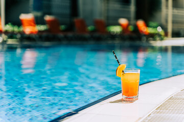 Tropical sparkling cocktail by the pool. The picture of glass with orange lemonade fruit cocktail standing near the poolside. Summer alcohol free drink by the hotel pool. Hello summer holiday vacation