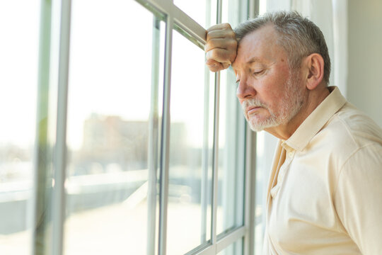 Sad tired lonely disappointed older senior man at home alone. Unhappy grandfather looking sadly outside window experiencing grief relative death bad news. Stressed elder man suffering from loneliness