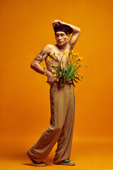 Full-length portrait of stylish, handsome, young man posing shirtless in beret and beige pants over yellow studio background. Concept of men's fashion, style, art, beauty, trends, summer inspiration