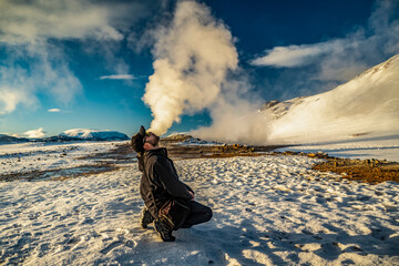 man imitates the release of smoke from his mouth with fumarole in the background at Hverir geothermal area in iceland