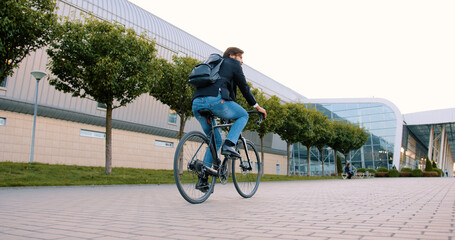 Good-looking stylish contented young bearded man riding a bicycle to his work on cobbled path near modern big bulding with glass windows,slow motion
