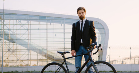 Good-looking stylish concentrated young businessman with well-groomed beard in formal clothes with backpack standing nearby his bike near modern panoramic building at dawn