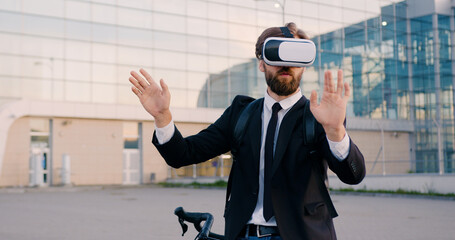 Front view of handsome confident purposeful young bearded office manr in augmented reality glasses which working on imaginary screen near modern glass building