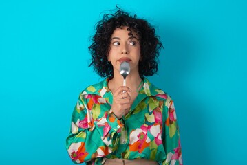 Very hungry Young arab woman wearing colorful shirt over blue background holding spoon into mouth...