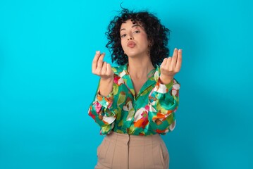 Young arab woman wearing colorful shirt over blue background doing money gesture with hands, asking...