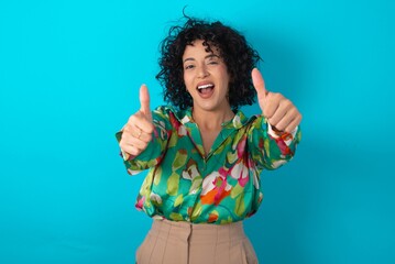 Young arab woman wearing colorful shirt over blue background approving doing positive gesture with...