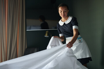 Young housekeeper or chambermaid in uniform changing blanket and other bedclothes on bed while...