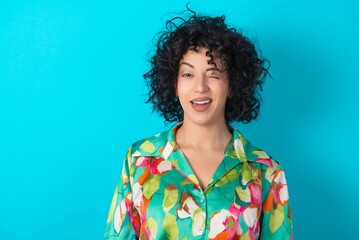 Coquettish young arab woman wearing colorful shirt over blue background smiling happily, blinking...