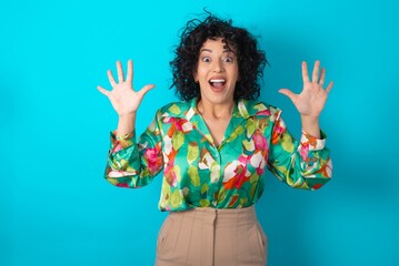 Optimistic young arab woman wearing colorful shirt over blue background raises palms from joy, happy to receive awesome present from someone, shouts loudly,