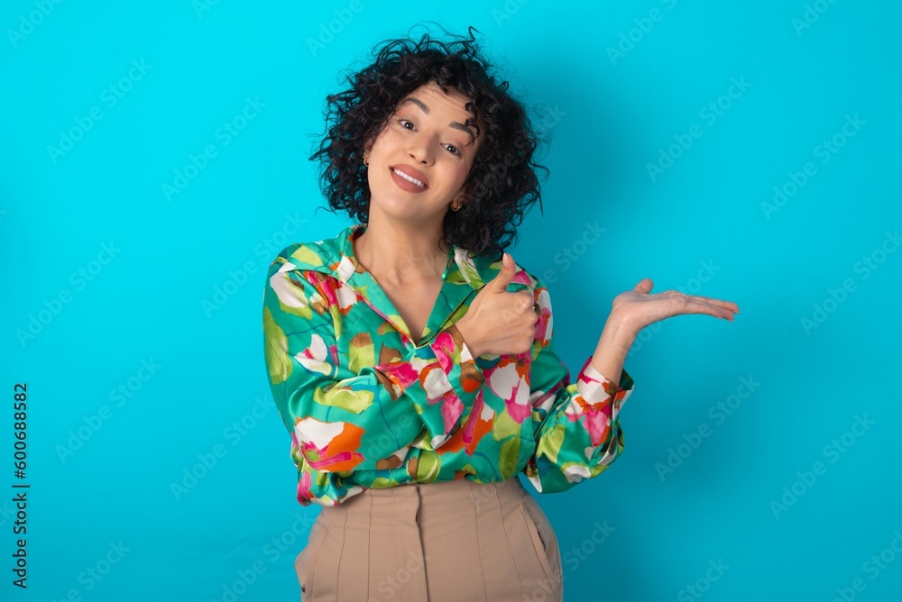 Poster young arab woman wearing colorful shirt over blue background Showing palm hand and doing ok gesture with thumbs up, smiling happy and cheerful.