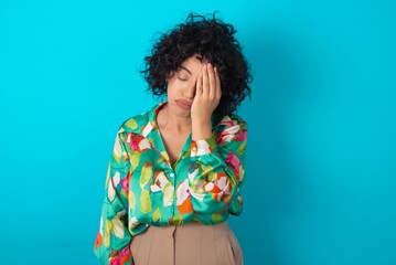 Tired overworked young arab woman wearing colorful shirt over blue background has sleepy...