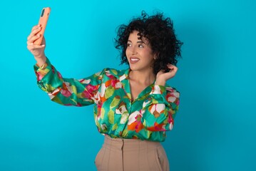 young arab woman wearing colorful shirt over blue background smiling and taking a selfie ready to...